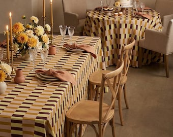 May include: A rectangular table and a round table covered in a geometric patterned tablecloth featuring alternating stripes of mustard yellow, dark brown, and light pink. Each table is set with crystal glassware, ceramic plates, and linen napkins.
