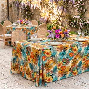 May include: A square tablecloth with a vibrant floral pattern in shades of yellow, orange, and turquoise. The table is set with white plates, glasses, and silverware. Wicker chairs surround the table, and a floral arrangement sits in the center.