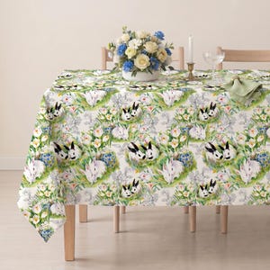 May include: A white tablecloth featuring a repeating pattern of watercolor-style rabbits and spring flowers in shades of green, yellow, and blue. The design includes small white and black bunnies nestled among crocus blooms and green foliage.