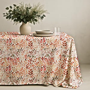May include: A rectangular tablecloth featuring an all-over pattern of small, watercolor-style leaves in shades of orange, red, brown, and muted green on a cream background. A beige ceramic vase with green foliage sits on the table surface.