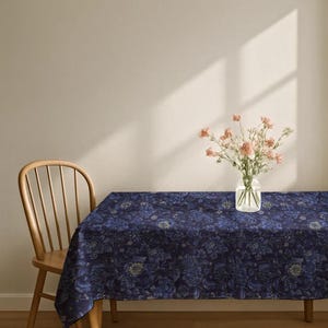 May include: A rectangular tablecloth featuring a dark navy blue fabric with an intricate, repeating floral pattern in shades of blue and muted gold. A clear glass vase containing a bouquet of light pink flowers sits on the tablecloth.