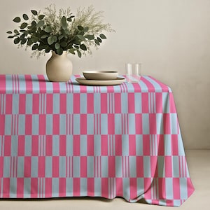 May include: A pink and light blue patterned tablecloth. The tablecloth has a repeating pattern of pink and light blue rectangles and vertical stripes. A vase with greenery, a bowl, and a plate are on the table.
