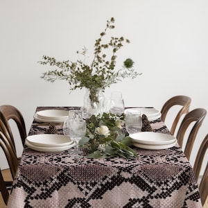 May include: A rectangular tablecloth featuring a repeating black and dusty pink snakeskin pattern. The table is set with white ceramic plates, clear ribbed drinking glasses, a glass vase filled with dried greenery, and a single white rose.