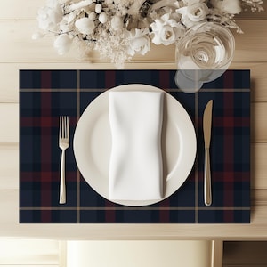 May include: A rectangular placemat featuring a dark navy blue, deep red, and thin gold plaid pattern. Centered on the placemat is a white ceramic dinner plate topped with a folded white napkin, a silver fork on the left, and a silver knife on the right.