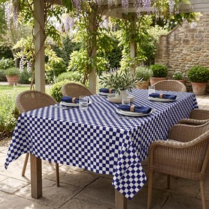 May include: A rectangular tablecloth featuring a blue and white checkered pattern draped over a wooden table. The table is set with white plates, navy blue napkins held by wooden rings, and clear glasses. Four woven wicker chairs surround the table.