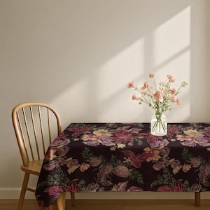 Può includere: Una tovaglia rettangolare con sfondo viola scuro e un motivo floreale ripetuto di fiori rosa, gialli e viola, foglie verdi e pigne. Un vaso di vetro trasparente contenente garofani rosa chiaro è posizionato sopra il tavolo.