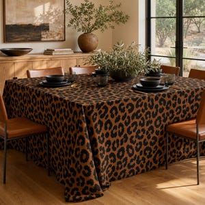 Könnte beinhalten: Eine quadratische Tischdecke mit einem sich wiederholenden schwarzen Leopardenmuster auf braunem Grund. Der Stoff liegt auf einem Esstisch, der mit schwarzen Tellern, Schüsseln und goldfarbenem Besteck gedeckt ist und ein markantes Bild bietet.