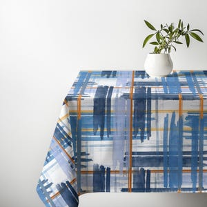 May include: A rectangular tablecloth featuring a plaid pattern with overlapping brushstrokes in shades of navy blue, light blue, and white, accented by thin, vertical and horizontal orange lines, draped over a surface with a white wall behind.