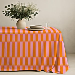 Puede incluir: Un mantel vibrante con un patrón repetitivo de rayas verticales naranjas y rosas, acentuado con pequeños rectángulos amarillos. Un jarrón con vegetación, un cuenco, un plato y un vaso de agua están sobre la mesa.