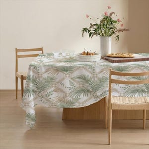 May include: A white tablecloth with a green and tan palm leaf pattern covers a wooden table. The table is set with a vase of flowers, a wooden cutting board, and a bowl of fruit. Two wooden chairs are visible.