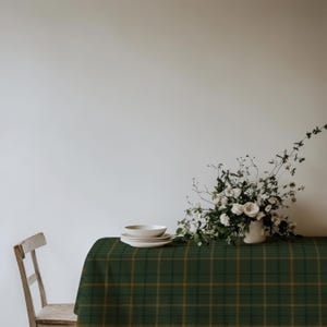 Op de afbeelding: Een donkergroen tafelkleed met een dun goud en zwart ruitpatroon over een rechthoekige tafel. Op de tafel staat een stapel van drie witte keramische borden en een witte keramische vaas gevuld met witte bloemen en groene takken.