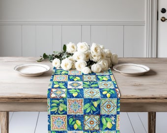 May include: A rectangular table runner featuring a repeating pattern of blue and white tiles interspersed with yellow lemons and green leaves. A bouquet of white roses sits on the runner between two white plates on a wooden table.