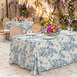 May include: A rectangular tablecloth featuring a blue and white toile pattern with illustrations of birds, trees, and foliage. The fabric is draped over a table set with white plates, clear glassware, and a colorful floral centerpiece.