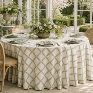 May include: A round dining table covered with a white tablecloth featuring a green leaf-patterned lattice design. The table is set with white plates, folded napkins, and clear glassware. A large bowl of white flowers sits in the center.