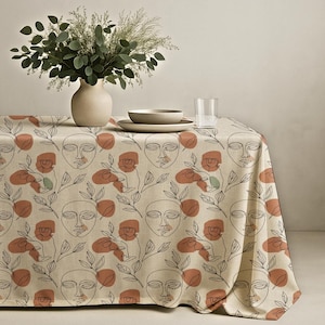 May include: A rectangular tablecloth featuring a repeating pattern of black line-art faces, leafy branches, and terracotta-colored abstract shapes on a beige background. The fabric drapes over a table surface, creating a textured, patterned display.