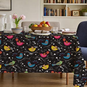 Könnte beinhalten: Eine schwarze Tischdecke mit einem sich wiederholenden Muster aus stilisierten Vögeln in Blau-, Gelb-, Rosa- und Grüntönen, durchsetzt mit weißen floralen und botanischen Strichzeichnungen. Der Stoff liegt auf einem rechteckigen Holztisch.