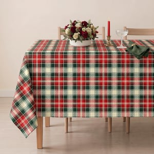 May include: A rectangular tablecloth featuring a classic plaid pattern in shades of red, dark green, and cream. The fabric drapes over a table, which is set with a white floral centerpiece in a bowl, a red taper candle, and a folded dark green napkin.