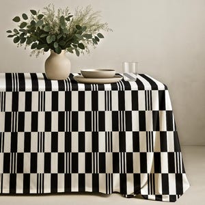 May include: A black and white checkered tablecloth with a vase of greenery, a bowl, and a plate. The tablecloth has a bold pattern of alternating black and white rectangles, with vertical white stripes. The vase is beige.