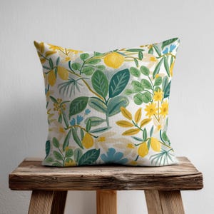 Könnte beinhalten: Quadratisches Kissen mit einem botanischen Zitronen- und Blumenmuster. Das Design zeigt gelbe Zitronen, grüne Blätter und blaue und gelbe Blumen auf weißem Hintergrund. Das Kissen liegt auf einem rustikalen Holzschemel.