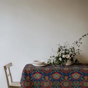 May include: A rectangular tablecloth featuring a vibrant, repeating mandala-inspired pattern in shades of deep blue, red, green, and yellow. A stack of white ceramic bowls and a white vase filled with white flowers sit on the table surface.
