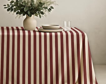 May include: A rectangular tablecloth featuring a vertical striped pattern in deep burgundy and cream. On the table sits a matte cream-colored vase filled with green eucalyptus branches, a shallow cream bowl, and a clear glass tumbler.