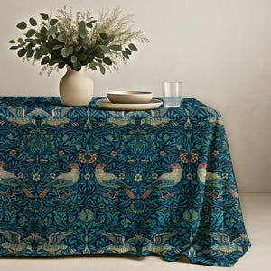 May include: A teal tablecloth featuring a repeating pattern of birds and floral designs. The birds are in tan, red, and teal. A vase with greenery, a bowl, a plate, and a glass of water are on the table, creating a dining setting.