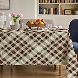 May include: A rectangular tablecloth featuring a diagonal plaid pattern in shades of cream, dark brown, and forest green. The fabric drapes over a dining table, covering the top and sides with a consistent geometric grid design.