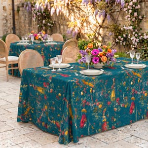 May include: A rectangular table covered with a teal tablecloth featuring a colorful floral and figure print. The table is set with white plates, clear glassware, and a central floral arrangement of pink, orange, and purple flowers.