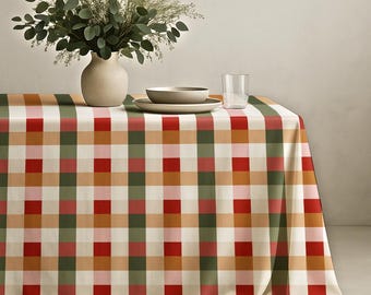 May include: A rectangular tablecloth featuring a large-scale plaid pattern in shades of red, dark green, light pink, and tan. On top sits a beige ceramic vase filled with green eucalyptus branches, a shallow beige bowl, and a clear glass tumbler.