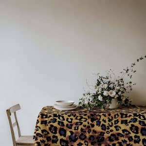 Op de afbeelding: Een rechthoekig tafelkleed met een opvallend luipaardpatroon in geel, zwart en bruin. Het tafelkleed ligt over een tafel met daarop een stapel van drie witte keramische kommen en een witte vaas gevuld met witte bloemen.