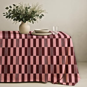 May include: A pink and burgundy checkered tablecloth with a vase of greenery, a bowl, and a plate. The tablecloth has a repeating pattern of pink and burgundy rectangles. The vase is beige, and the bowl and plate are light gray.