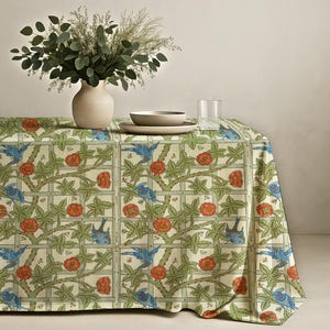 Peut inclure: Une nappe crème avec un motif répété d'oiseaux bleus, de roses rouges et de vignes vertes. La nappe est posée sur une table, avec un vase de verdure, un bol, une assiette et un verre d'eau.