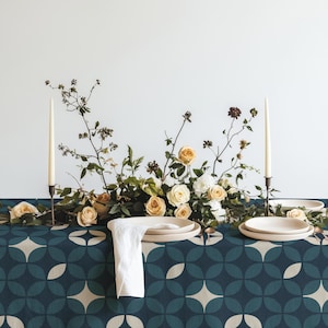 Könnte beinhalten: Eine dunkel-türkisfarbene und cremefarbene Tischdecke mit geometrischem Muster und einem Blumenarrangement aus cremefarbenen und pfirsichfarbenen Rosen mit grünem Blattwerk. Zwei hohe cremefarbene Stabkerzen in dunklen Haltern flankieren das Gesteck.