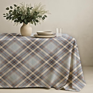 May include: A rectangular tablecloth featuring a diagonal plaid pattern in shades of light blue, brown, and cream. The fabric drapes over a table surface, providing a structured textile base for a ceramic vase with greenery, a bowl, and a glass.
