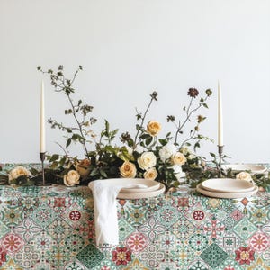 May include: A table setting featuring a tablecloth with a multi-colored geometric tile pattern in shades of green, red, and yellow. The arrangement includes two tall white taper candles, a central floral centerpiece, and white ceramic plates.