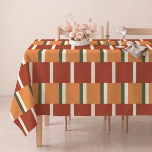 Könnte beinhalten: Eine rechteckige Tischdecke mit einem geometrischen Muster aus abwechselnden vertikalen Blöcken in gebranntem Orange, Terrakotta und Creme, die durch schmale dunkelgrüne Streifen getrennt sind. Der Stoff liegt auf einem Tisch und bildet ein strukturiertes Muster.