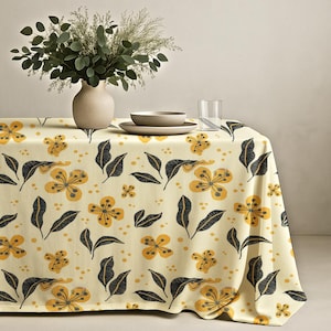 May include: A cream-colored tablecloth with a repeating pattern of yellow flowers and dark gray leaves. A vase with greenery, a bowl, a plate, and a glass of water are on the table. The tablecloth is draped over the table.