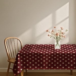 May include: A rectangular tablecloth featuring a deep burgundy background with a repeating pattern of small, light cream-colored polka dots. A clear glass vase containing a bouquet of light pink carnations sits on the center of the table.