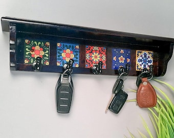 May include: A dark brown wooden key holder with a shelf and five hooks. The key holder features colorful, patterned tiles with floral designs. Keys hang from the hooks. The key holder is mounted on a gray wall.