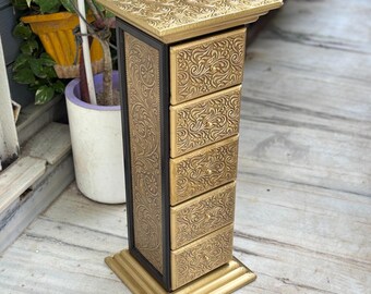 May include: A gold-colored, ornate jewelry chest with five drawers and a square top. The chest features intricate floral patterns and a black frame. The base is also gold, and the chest stands tall, perfect for storing small items.