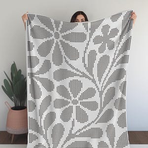 Könnte beinhalten: Eine weiße Decke mit einem schwarzen Blumenmuster. Das Design zeigt große stilisierte Blumen und Blätter, die mit einem pixeligen Effekt erstellt wurden. Die Decke wird hochgehalten und zeigt das gesamte Muster.