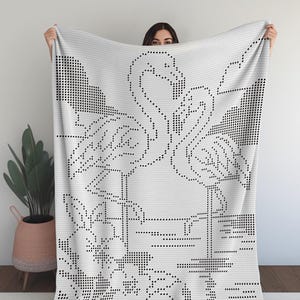 Op de afbeelding: Een witte deken met een zwart stippenpatroon met twee flamingo's, bloemen en bladeren. De deken wordt omhoog gehouden, waarbij het gedetailleerde patroon wordt getoond. Het ontwerp doet denken aan kruissteek of pixelkunst.