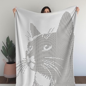 May include: A white blanket with a black pixelated cat design. The cat's face is the main feature, with whiskers and a detailed outline. The blanket is made of a soft material, ideal for cosy nights.