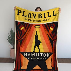Puede incluir: Una manta amarilla, roja y negra con el diseño del programa del musical "Hamilton". La manta muestra las palabras "PLAYBILL" y "RICHARD RODGERS THEATRE" en la parte superior, con el título del musical y el logotipo de la estrella debajo.