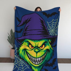 May include: A blue blanket featuring a pixelated image of a green creature with a purple witch hat, a wide grin, and red eyes. Black bats and a spiderweb design are in the background. The blanket is held up by a person.