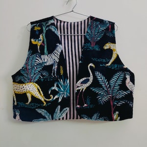 Op de afbeelding: Een mouwloos vest met een zwarte jungleprint met zebra's, luipaarden en vogels. De binnenvoering van het vest heeft verticale roze en witte strepen. Het vest hangt aan een metalen hanger.