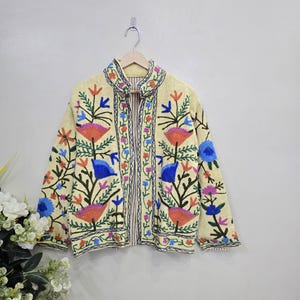 May include: A cream-colored jacket featuring vibrant, multi-colored floral embroidery in shades of orange, blue, and pink. The jacket has a high collar, long sleeves, and a vertical striped lining visible at the front opening, hanging on a hanger.