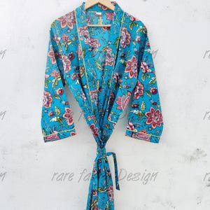 May include: A turquoise robe featuring a vibrant floral pattern in shades of pink, red, and green. The garment has long sleeves, a wrap-front design with a matching tie belt, and thin yellow piping along the edges. The text rare fabric Design is visible.