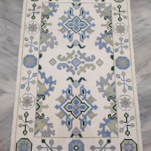 May include: Rectangular off-white rug with a geometric pattern in shades of blue, gray, and green. The rug features a central design and a border with repeating floral and geometric motifs. The rug has a fringe on the short ends.