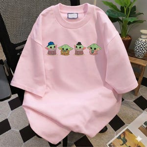 Pu&ograve; includere: Una t-shirt a maniche corte color rosa chiaro con una stampa grafica di quattro piccole creature verdi dalle grandi orecchie. Le creature sono raffigurate in fila, la prima indossa un berretto blu, la terza un cappello nero e la seconda tiene una tazza.